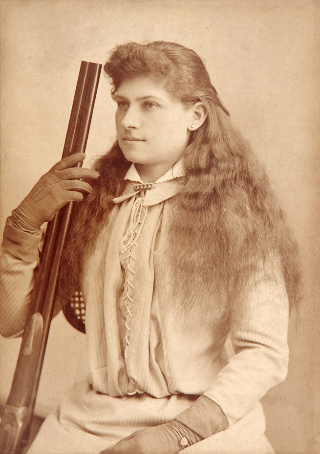Sepia image of Annie Oakley sitting, angled to her right with perfect posture, and holding a shotgun barrel up in her gloved right hand with her left hand in her lap
